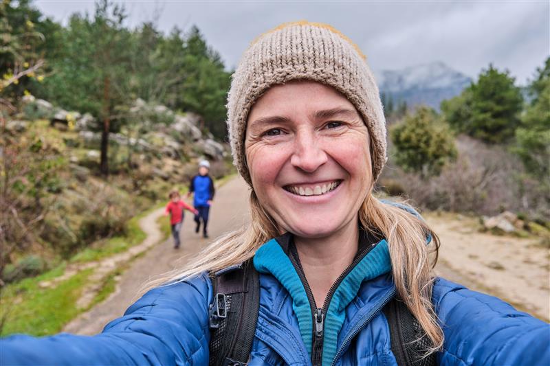 Woman taking a selfie on a hiking trail with a child and another person in the background.