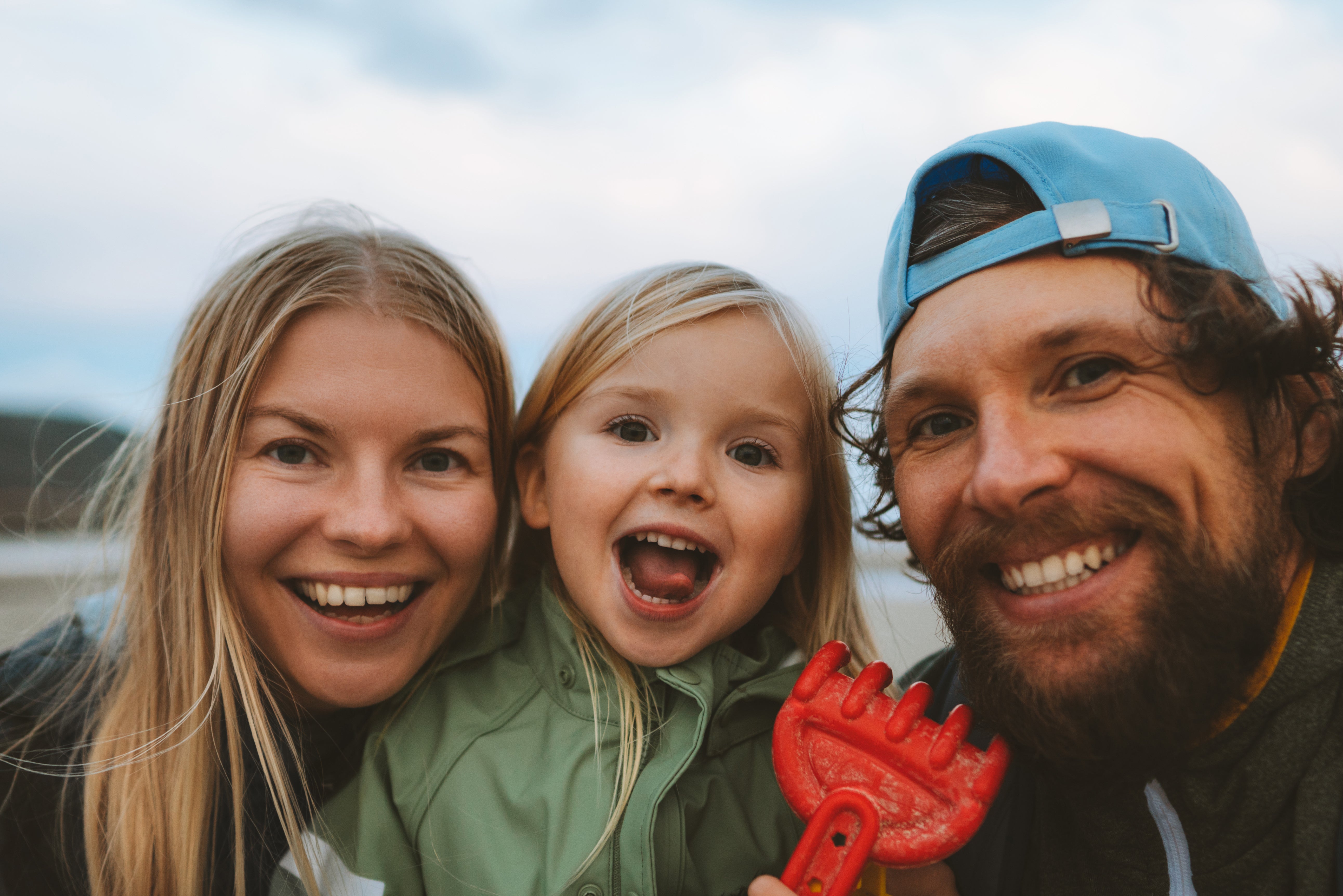 Family of three outdoors with a red object, smiling at the camera.