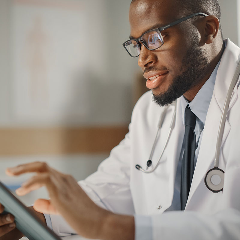 Happy and Smiling African American Male Doctor Wearing White Coat Working on Tablet Computer at His Office. Medical Health Care Professional Working with Test Results, Patient Treatment Planning.