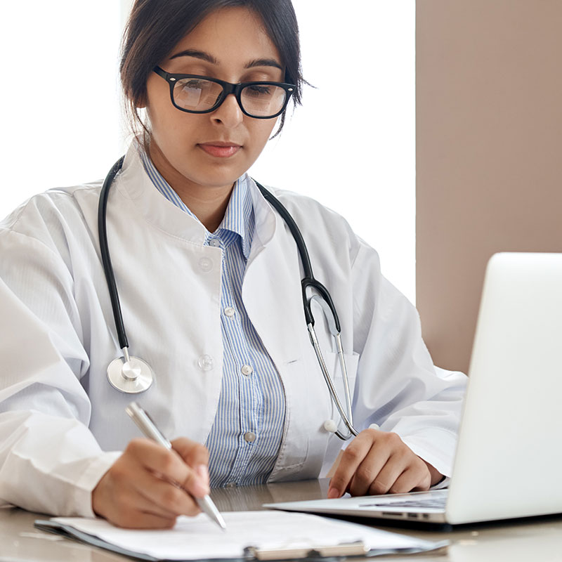 Indian ethnic female doctor physician gp wearing glasses, white coat stethoscope writing filling medical form watching online medical webinar seminar training working sitting with laptop at workplace.